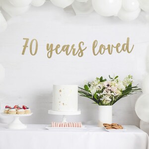 May include: A white table with a white tablecloth, a white cake with gold flecks, a bouquet of white flowers in a white vase, and a banner that reads "70 years loved".