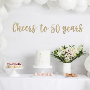 May include: A white table with a white tablecloth set with a white cake, cupcakes, cookies, and macarons. A bouquet of white flowers in a white vase is on the table. A gold glitter banner reads "Cheers to 50 years". White balloons are in the background.