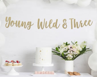 Może przedstawiać: Scena imprezy z białym obrusem, ciastem i balonami. Baner głosi "Young Wild & Three" złotym pismem. Białe ciasto ze złotymi akcentami stoi na cokole, z babeczkami i ciasteczkami w pobliżu. Bukiet kwiatów znajduje się w białym wazonie.