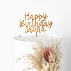 May include: A white cake with a wooden cake topper that says "Happy Birthday Laura" and a bouquet of dried flowers.