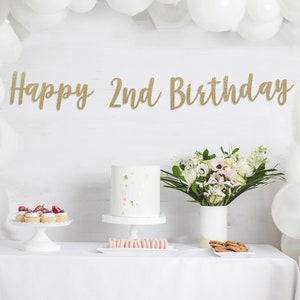 Op de afbeelding: Een witte tafel met een wit tafelkleed, een witte taart met gouden sprinkles, cupcakes met bessen, een boeket witte bloemen en een gouden glitterbanner met de tekst "Happy 2nd Birthday".