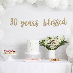 May include: A white table with a white tablecloth, a white cake with gold flecks, a white cake stand, a plate of cupcakes, a plate of cookies, a vase with white flowers, and a gold glitter banner that says "90 years blessed".