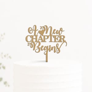 May include: A gold-colored cake topper featuring the text A New Chapter Begins in a decorative script font, positioned on top of a plain white cylindrical cake that rests on a black pedestal stand, with a small heart icon above the word New.