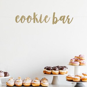 May include: A white table with four tiers of donuts. The top tier has a stack of pink and white donuts. The second tier has a ring of chocolate-covered donuts. The third tier has a row of pink-frosted donuts. The bottom tier has a row of plain donuts. A gold glitter banner that says "cookie bar" hangs above the table.
