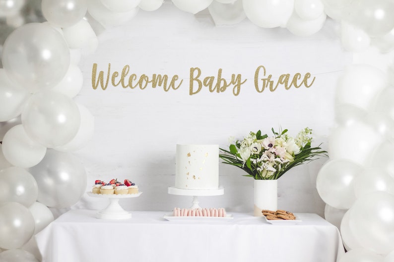 May include: A white table with a white tablecloth, a white cake, cupcakes with berries, and a bouquet of white flowers. A gold glitter banner reads "Welcome Baby Grace".