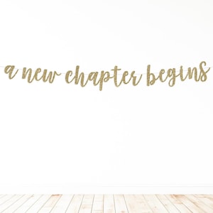May include: A gold glitter banner with the phrase "a new chapter begins" strung across a thin string. The banner is displayed against a white wall with a light wood floor.