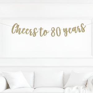 May include: A gold glitter banner featuring the text Cheers to 80 years in a cursive script font. The banner is suspended on a thin white string, with each letter cut out individually and connected to form the celebratory phrase for an event.