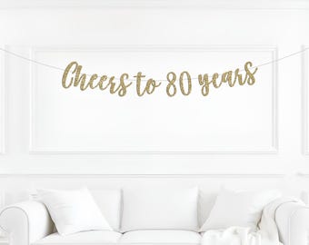 May include: A gold glitter banner featuring the text Cheers to 80 years in a cursive script font. The banner is suspended on a thin white string, with each letter cut out individually and connected to form the celebratory phrase for an event.