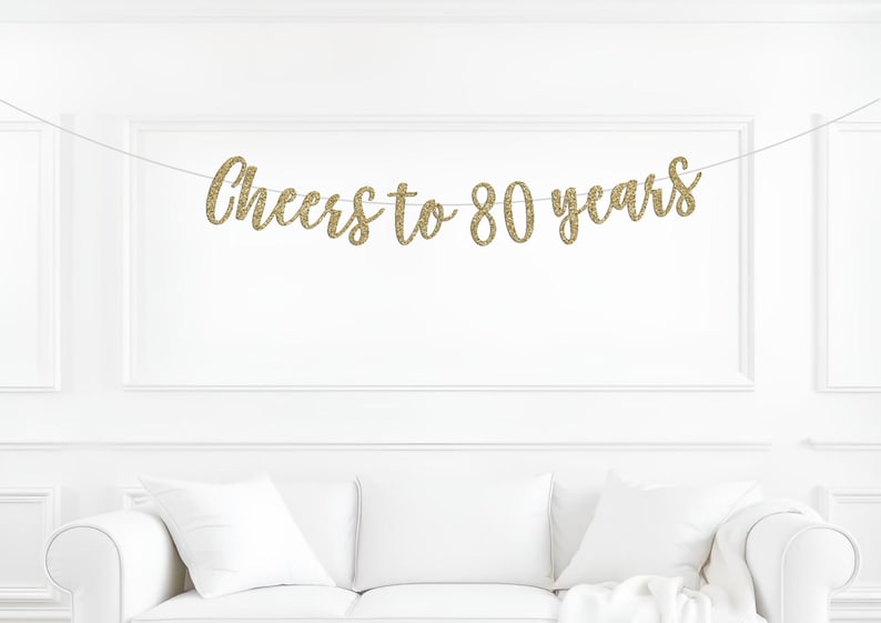 May include: A gold glitter banner featuring the text Cheers to 80 years in a cursive script font. The banner is suspended on a thin white string, with each letter cut out individually and connected to form the celebratory phrase for an event.