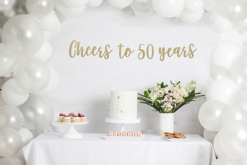 May include: A white table with a white tablecloth set with a white cake, cupcakes, cookies, and macarons. A bouquet of white flowers in a white vase is on the table. A gold glitter banner reads "Cheers to 50 years". White balloons are in the background.