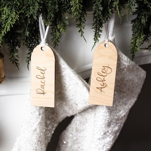 May include: Two wooden gift tags with the names "Rachel" and "Ashley" engraved in a cursive font. The tags are hanging from white ribbons, with a white sequined stocking in the background. Greenery is visible at the top.