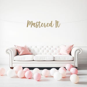 May include: A white tufted sofa with pink pillows and scattered pink and white balloons. A gold glitter banner that reads "Mastered It" hangs above the sofa, creating a celebratory atmosphere.