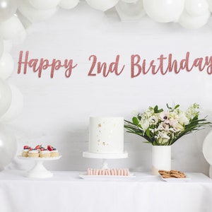 Op de afbeelding: Een witte tafel met een wit tafelkleed gedekt voor een verjaardagsfeestje. Er is een witte taart met gouden accenten, cupcakes met bessen, macarons en koekjes. Een boeket witte bloemen in een witte vaas staat op de tafel. Een banner met roze gouden glitterletters luidt "Happy 2nd Birthday".
