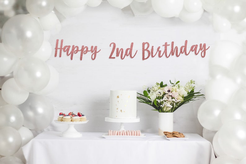 Op de afbeelding: Een witte tafel met een wit tafelkleed gedekt voor een verjaardagsfeestje. Er is een witte taart met gouden accenten, cupcakes met bessen, macarons en koekjes. Een boeket witte bloemen in een witte vaas staat op de tafel. Een banner met roze gouden glitterletters luidt "Happy 2nd Birthday".
