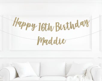 May include: A gold glitter banner that reads "Happy 16th Birthday Maddie" is strung across a white wall. The banner is above a white couch with white pillows. The text is in a cursive font.