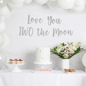 May include: A white table is set for a celebration, featuring a cake with gold accents, cupcakes, macarons, and cookies. The backdrop includes white balloons and a banner that reads "Love You TWO the Moon."