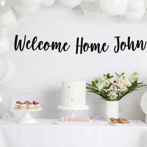 May include: A white table with a white tablecloth, decorated with white balloons, a white cake with gold sprinkles, a bouquet of white flowers, and a sign that says "Welcome Home John".