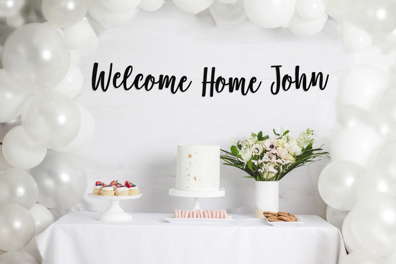 May include: A white table with a white tablecloth, decorated with white balloons, a white cake with gold sprinkles, a bouquet of white flowers, and a sign that says "Welcome Home John".