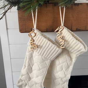 May include: Two white knitted stockings with wooden name tags that say "Mom" and "Dad". The stockings are hanging from a wooden mantelpiece.