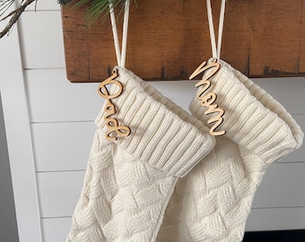 May include: Two white knitted stockings with wooden name tags that say "Mom" and "Dad". The stockings are hanging from a wooden mantelpiece.