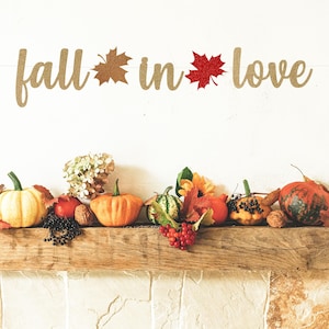 May include: A wooden sign with the text "fall in love" decorated with fall leaves. The sign is displayed on a wooden mantel with a variety of fall decorations, including pumpkins, gourds, and berries.