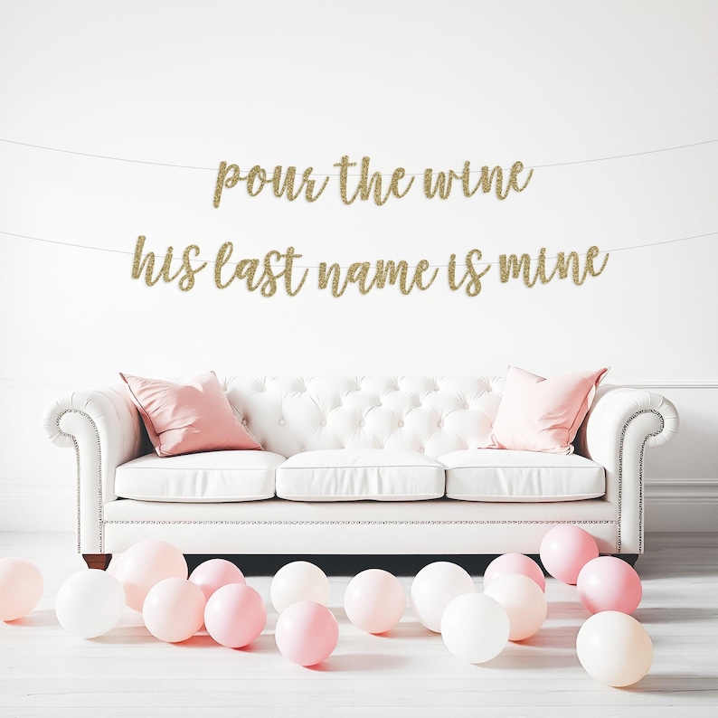 May include: Gold glitter banner with the text "pour the wine his last name is mine" hanging on a white wall above a white sofa with pink cushions and pink and white balloons on the floor.