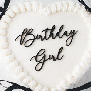 May include: White heart-shaped cake with black frosting and a black ribbon. The cake is decorated with the words "Birthday Girl" in black lettering.