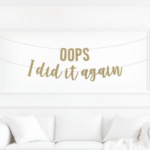 Peut inclure: Une bannière à paillettes dorées affichant le texte OOPS I did it again, suspendue par deux fines ficelles blanches. Les lettres sont disposées sur deux lignes avec OOPS en haut et I did it again en bas, le tout avec une finition scintillante.