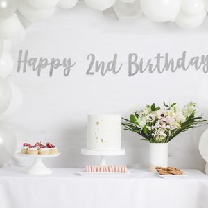 Op de afbeelding: Een witte tafel met een wit tafelkleed, een witte taart met gouden sprinkles, cupcakes met rode bessen, roze macarons en een boeket witte bloemen in een witte vaas. Een zilveren glitterbanner met de tekst "Happy 2nd Birthday".
