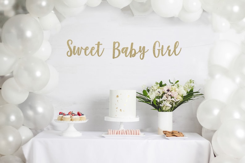 May include: A white table with a white tablecloth, a white cake with gold flecks, cupcakes with strawberries, a bouquet of white flowers in a white vase, and a banner that reads 'Sweet Baby Girl' in gold glitter letters.