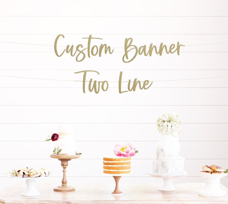 以下が含まれることがあります： 白い木製の背景に、「Custom Banner Two Line」という文字が書かれた金色のグリッターバナーが吊り下げられています。バナーの前に、ケーキスタンドに3つのケーキが飾られています。