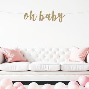 May include: A gold glitter "Oh Baby" banner hangs above a white sofa with pink cushions. Pink and white balloons are scattered on the floor in front of the sofa.
