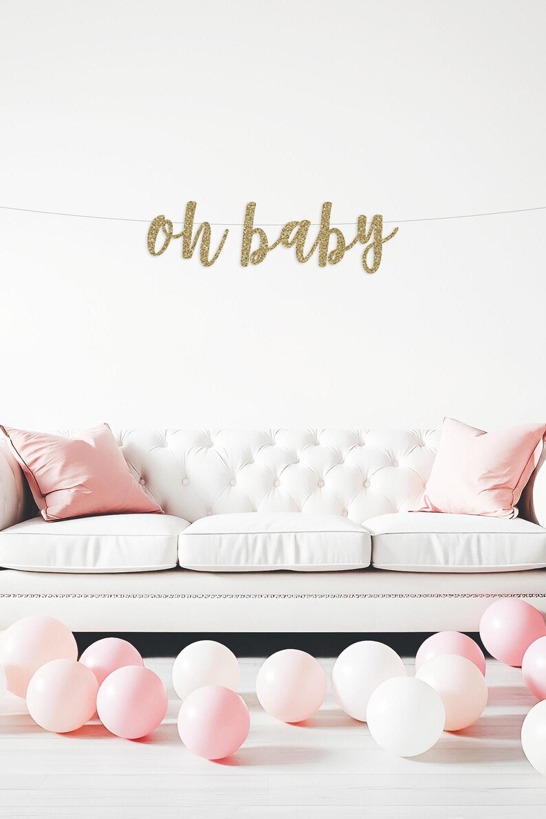 May include: A gold glitter "Oh Baby" banner hangs above a white sofa with pink cushions. Pink and white balloons are scattered on the floor in front of the sofa.