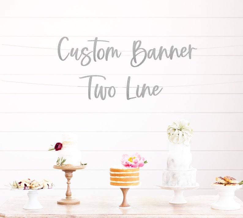 以下が含まれることがあります： 白い木製の壁に、「Custom Banner Two Line」という文字が書かれた銀色のグリッターバナーが掛けられています。木製のテーブルの上に、異なる装飾が施された3つのケーキがケーキスタンドに飾られています。