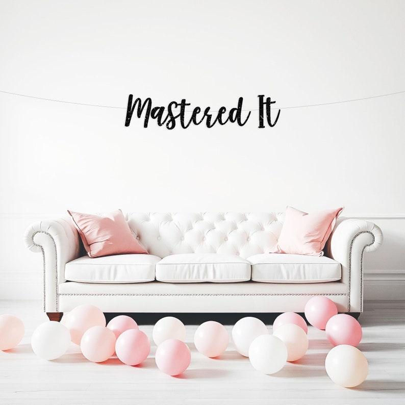 May include: A white tufted sofa with pink pillows and scattered balloons. A banner with the words "Mastered It" hangs above the sofa, creating a celebratory atmosphere. The balloons are a mix of white and light pink.
