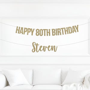 May include: A gold glitter banner featuring the text HAPPY 80TH BIRTHDAY on the top line and the name Steven on the bottom line, both suspended on thin white strings against a plain white wall above a white sofa with white cushions.