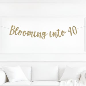 May include: A gold glitter banner that reads "Blooming into 40" is strung across a white wall. The banner is above a white couch with white pillows. The banner is a festive decoration for a milestone birthday.