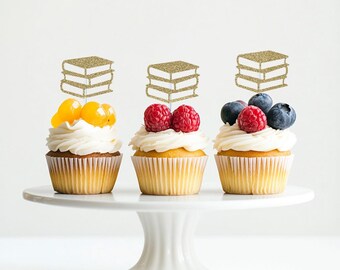 May include: Three cupcakes decorated with gold glitter book toppers, each cupcake is topped with fresh fruit, two cupcakes have raspberries and blueberries, and one cupcake has yellow fruit.