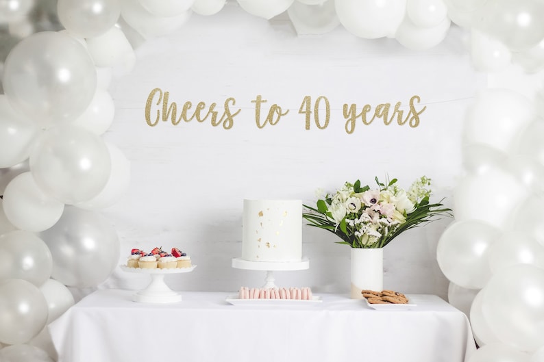 May include: A white table with a white tablecloth set for a party. There is a white cake with gold flecks, cupcakes with red berries, macarons, and cookies on the table. A bouquet of white flowers in a white vase is on the table. A gold glitter banner reads "Cheers to 40 years". White balloons are in the background.