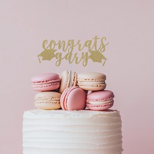 May include: A white cake with a gold glitter cake topper that says "congrats gary" with graduation caps on either side. The cake is decorated with pink and beige macarons.