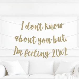May include: A gold glitter banner with the text "I don't know about you but I'm feeling 20x2" strung across a white wall. A white couch with pillows is below the banner.