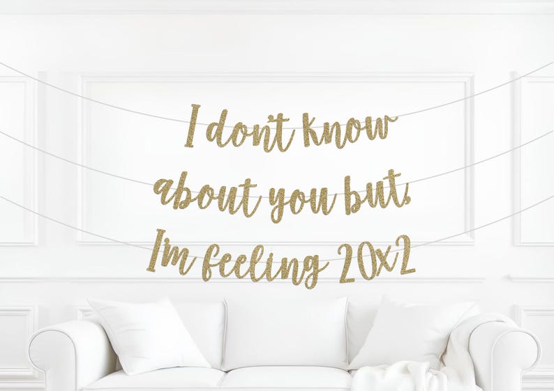 May include: A gold glitter banner with the text "I don't know about you but I'm feeling 20x2" strung across a white wall. A white couch with pillows is below the banner.