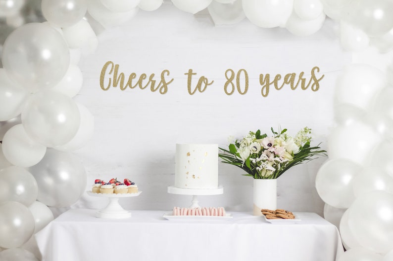 May include: A white table with a white tablecloth. On the table are cupcakes with red berries, a white cake with gold flakes, pink macarons, and cookies. A bouquet of white flowers in a white vase is on the table. A white banner with gold glitter letters reads 'Cheers to 80 years'. A white balloon arch is in the background.
