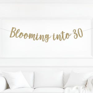 May include: A gold glitter banner that reads "Blooming into 30" is strung across a white wall. The banner is above a white couch with white pillows. The text is in a cursive font.