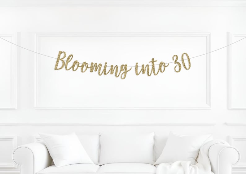 May include: A gold glitter banner that reads "Blooming into 30" is strung across a white wall. The banner is above a white couch with white pillows. The text is in a cursive font.