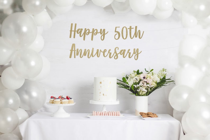 May include: A white table with a white tablecloth set for a 50th anniversary party. There is a white cake with gold flecks, cupcakes with berries, and a bouquet of white flowers in a white vase. The background is a white wall with a gold glitter banner that reads "Happy 50th Anniversary".