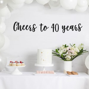 May include: A white table with a white tablecloth, decorated with white balloons, a white cake with gold accents, a bouquet of white and pink flowers in a white vase, and a black banner that reads "Cheers to 40 years".
