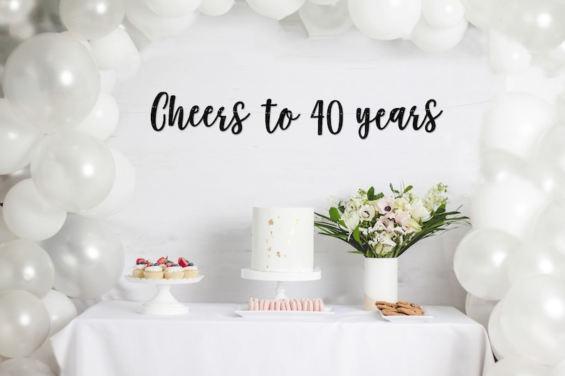 May include: A white table with a white tablecloth, decorated with white balloons, a white cake with gold accents, a bouquet of white and pink flowers in a white vase, and a black banner that reads "Cheers to 40 years".