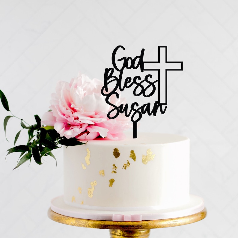 May include: Black acrylic cake topper with the text "God Bless Susan" and a cross. The topper is on a white cake with gold flecks.