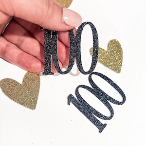 May include: Black glittery number "100" cutouts with a hand holding one. Gold glitter heart shapes are scattered around the numbers on a white background. The cutouts are likely for a milestone celebration.